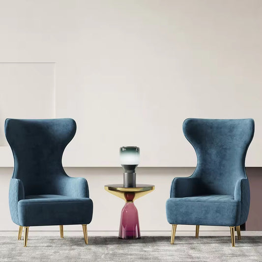 Two high back luxury accent chairs with a blue upholstery, gold legs, and a round table between them, set against a plain background.
